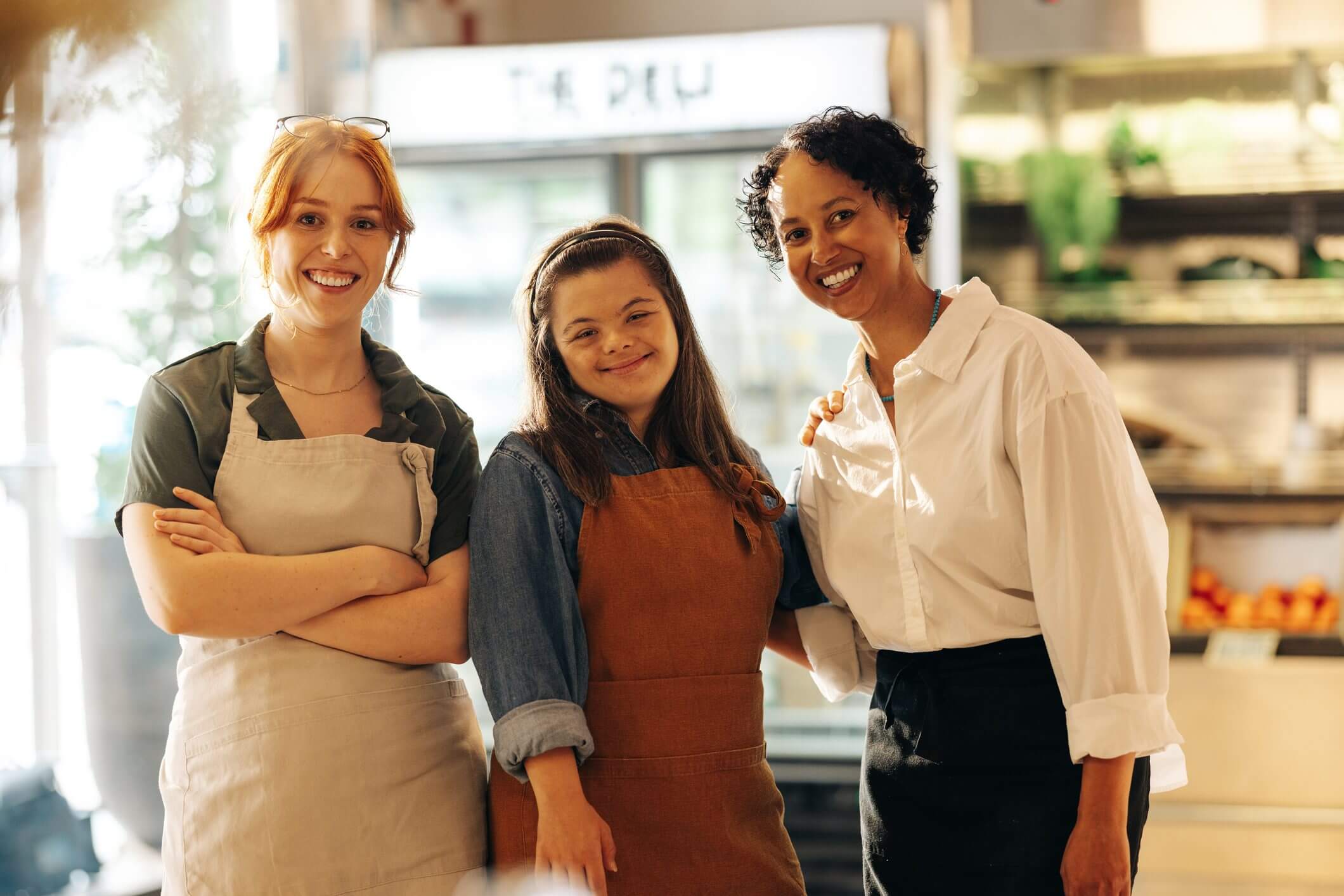 Three women smiling, two wearing aprons, posing in a cafe.