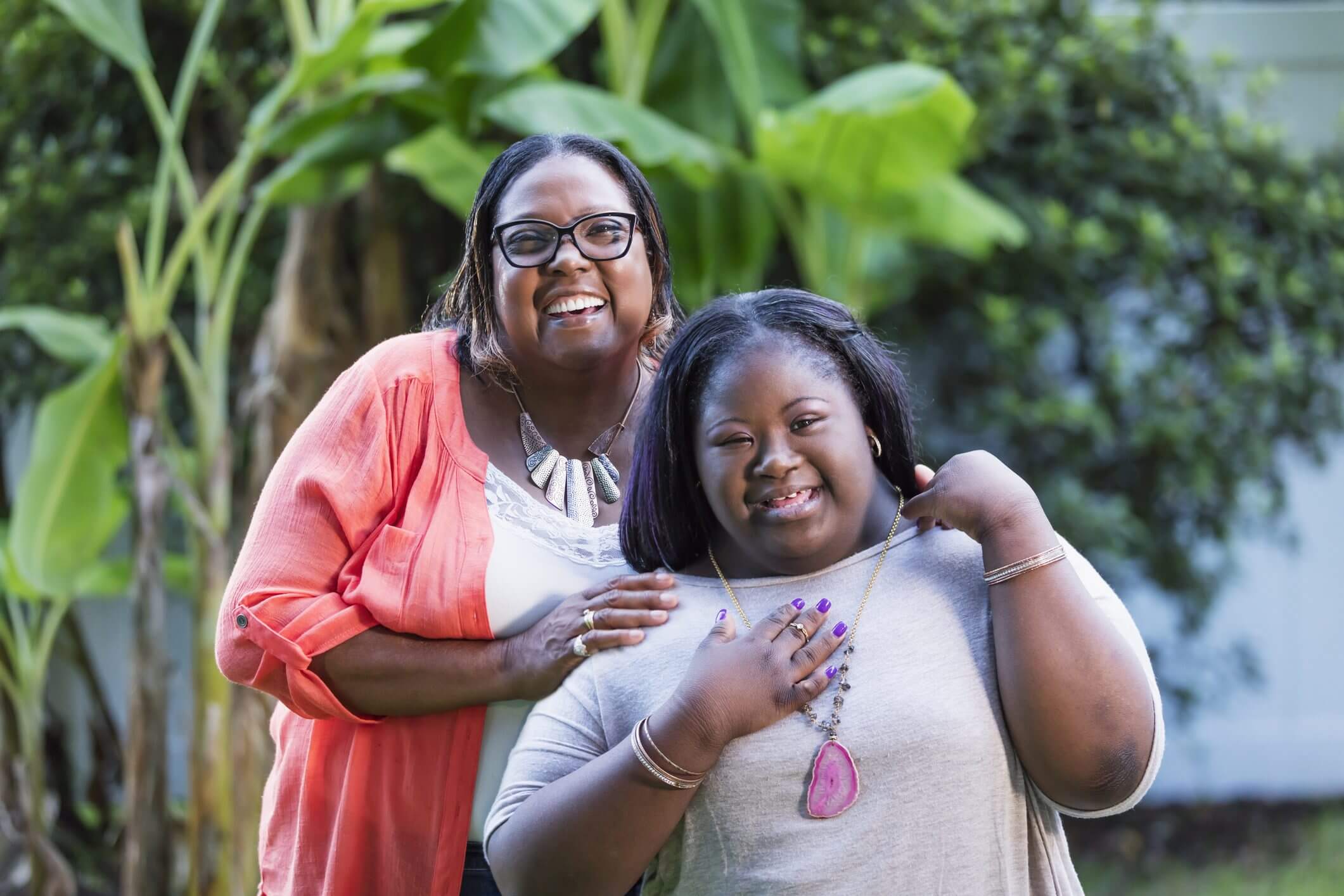 Two women smiling warmly outdoors with greenery in the background.