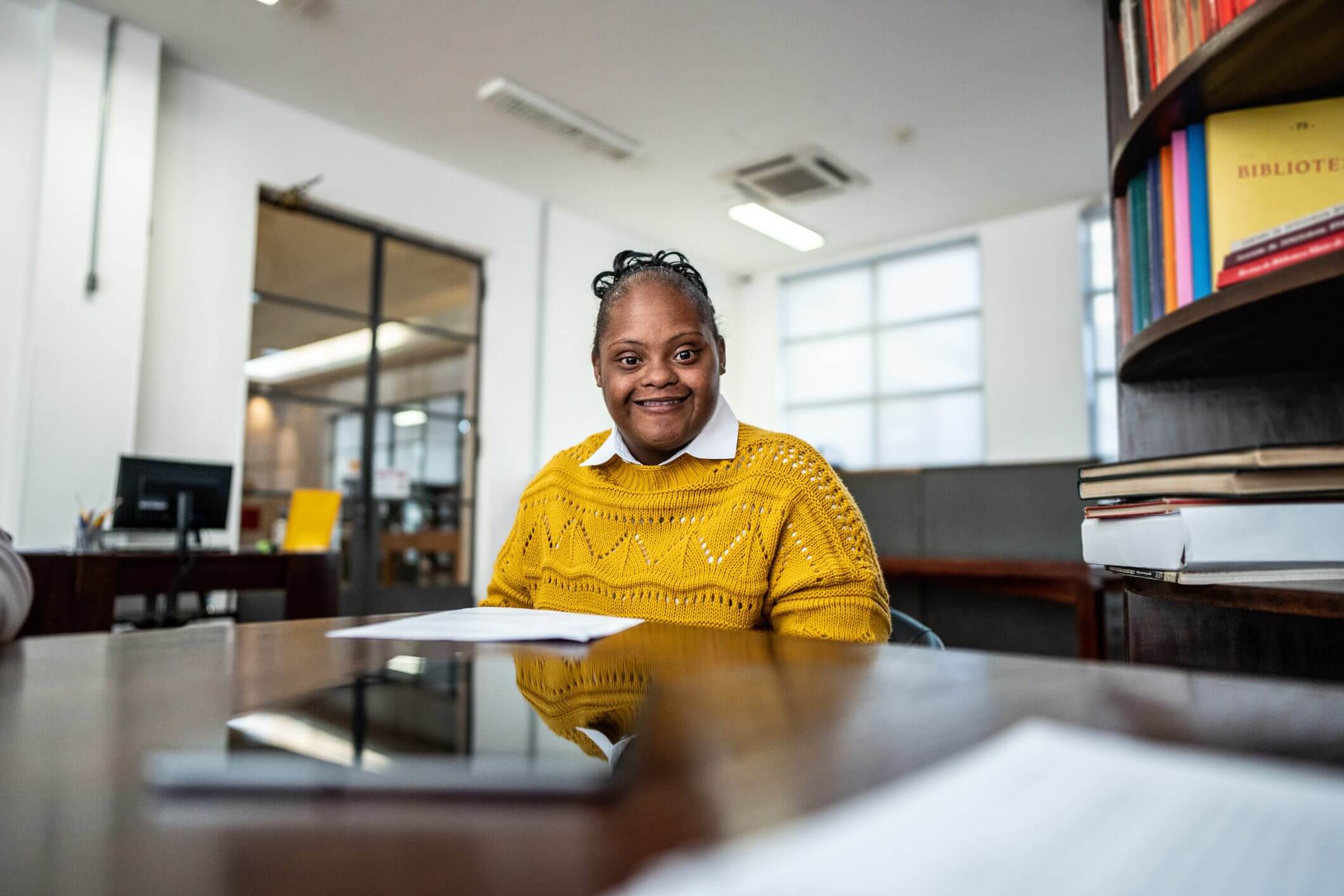 Smiling woman in a yellow sweater sitting at a desk in a bright office.